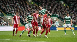 Soccer players in green and white hooped shirts and red and white striped shirts contest a header in a packed stadium