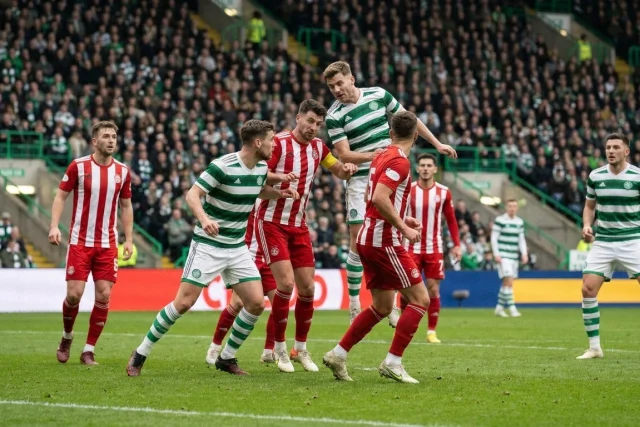 Soccer players in green and white hooped shirts and red and white striped shirts contest a header in a packed stadium
